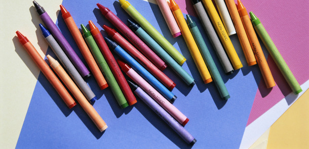 Mehrere Wachsmalstifte liegen nebeneinander auf buntem Papier auf einem Tisch.