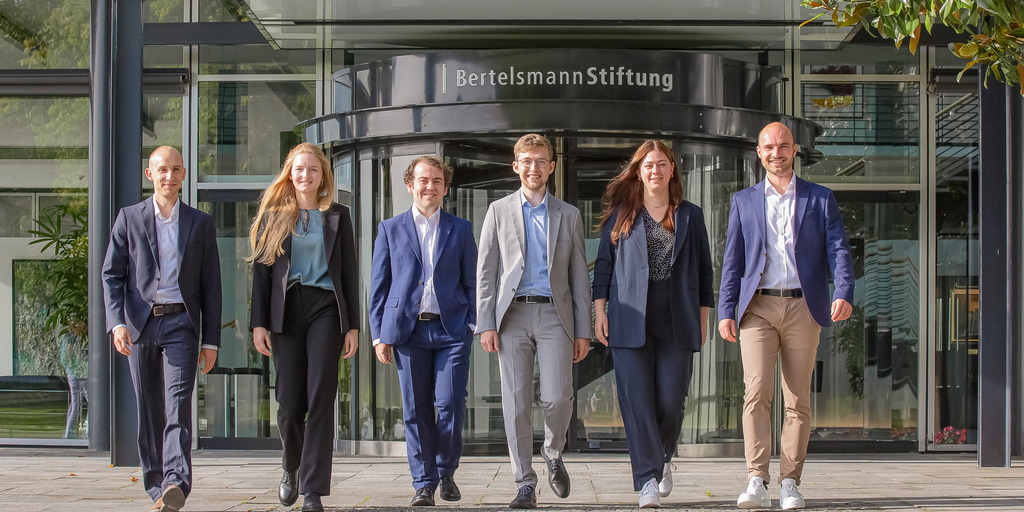 Die sechs Junior Professionals der Bertelsmann Stiftung des Jahrgangs 2023 - Jakob Weber, Anne Meisiek, Etienne Höra, Leander Berner, Lena Budach und Otto Meyer zu Schwabedissen - gehen gemeinsam den Weg vor dem Eingang zur Bertelsmann Stiftung hinunter und blicken in die Kamera.
