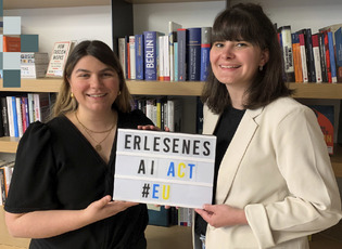 Teresa Staiger und Asena Soydas mit einer Light-Box zu sehen. Sie stehen vor einem Bücherregal und die Lightbox hat den Schriftzug: Erlesenes AI Act