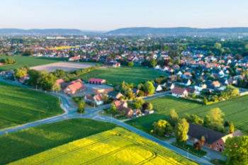 Das Foto zeigt eine Panoramaaufnahme einer kleinen Stadt, welche im Hintergrund zu sehen ist. Im Vordergrund liegen Felder und kleine Wälder. Der Himmel ist hellblau mit leichten Wolken.