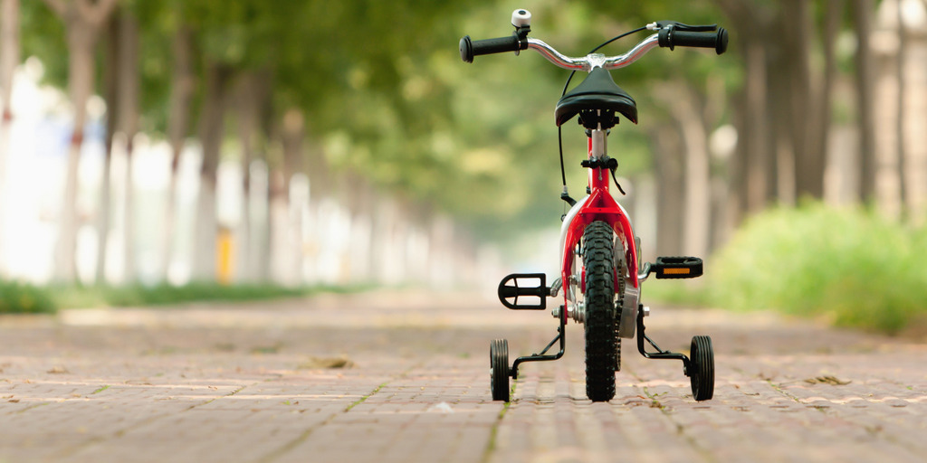 Ein Kinderfahrrad mit Stützrädern steht verlassen auf einem von Bäumen gesäumten Weg.