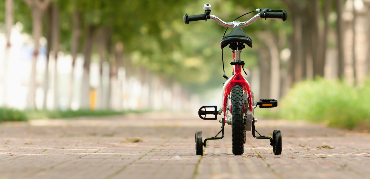 (© Shutterstock / L.F) Ein Kinderfahrrad mit Stützrädern steht verlassen auf einem von Bäumen gesäumten Weg.