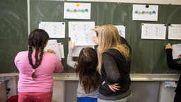 Zwei Schülerinnen stehen an der Tafel und bekommen von ihrer Lehrerin etwas erklärt.