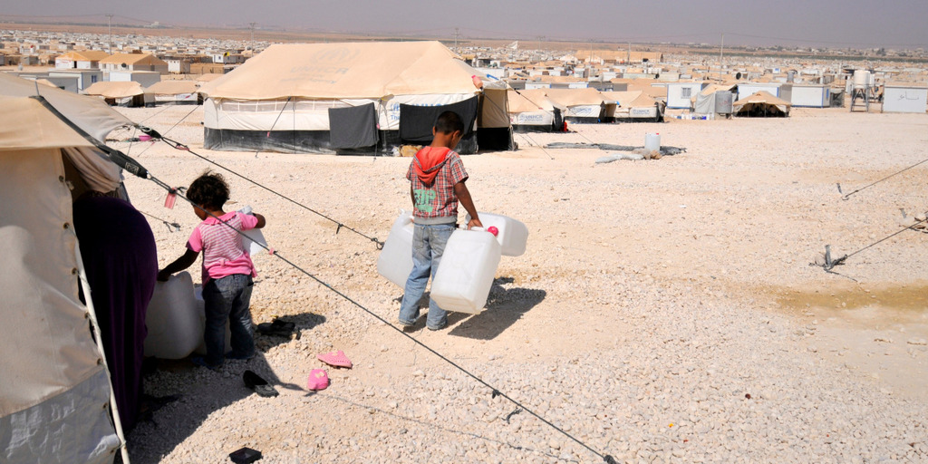 [Translate to English:] Blick auf das jordanische Flüchtlingslager Zaatari. Zu sehen sind hunderte befestigte Unterkunfszelte, die bis zum Horizont reichen. Im Vordergrund stehen zwei Kinder mit Wasserkanistern, die der Kamera den Rücken zugedreht haben