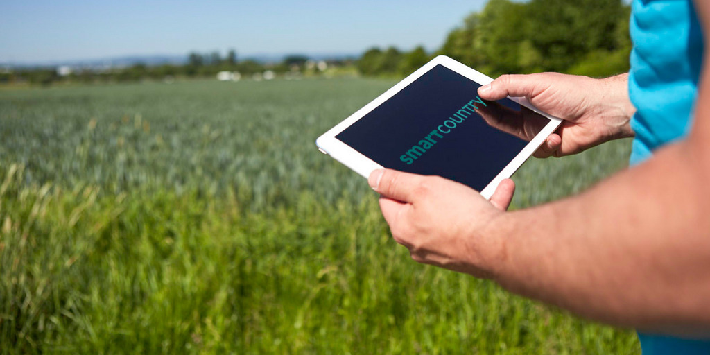 vor einer Grünen Wiese halten zwei Hände einen Tablet-PC, auf dem Display steht "Smart Country"