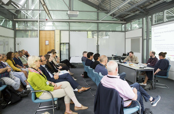Das Foto zeigt einen Workshopraum. Vorne stehen die Referent:innen. Seitlich links die Stuhlreihen mit den Teilnehmenden des Workshops.