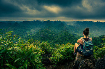 Eine Wanderin sitzt auf einem Stein auf einem Hügel und blickt auf eine weitläufige, dicht bewachsene Dschungellandschaft.