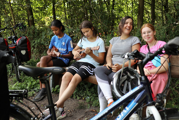 Schülerinnen und eine Lehrerinn machen Pause auf eine Radtour. Ein Mädchen spielt Ukulele