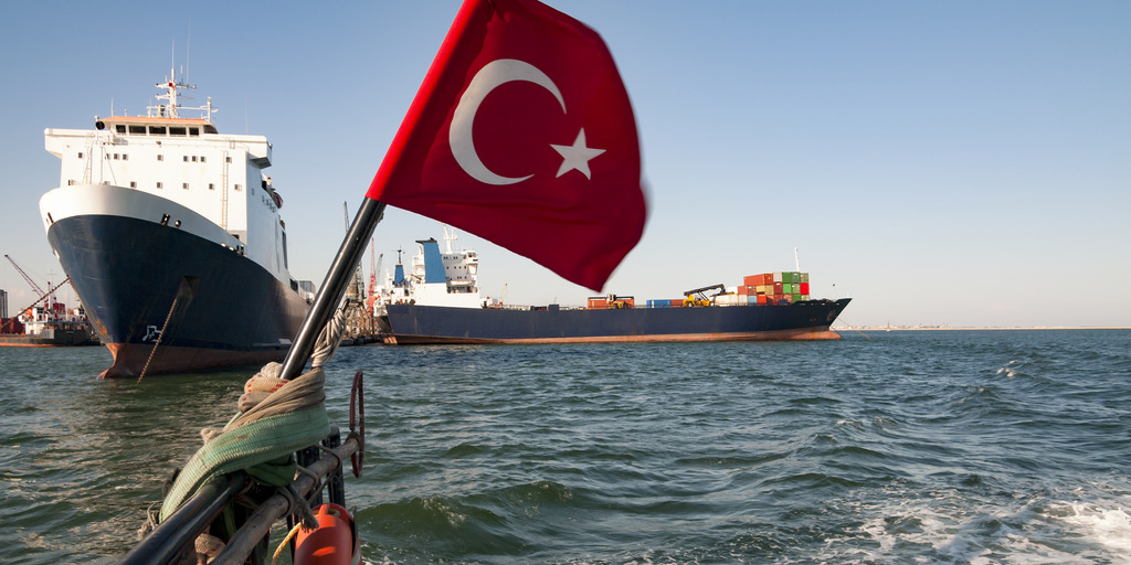 Ein Boot mit türkischer Flagge treibt im Meer. Im Hintergrund sind zwei Containerschiffe und ein Schiff mit Verladekran zu sehen.