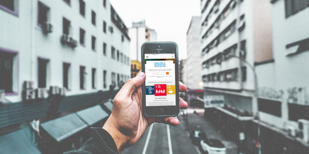 Im Vordergrund hält eine Hand ein Handy. Auf dem Handy ist die Seite des SDG-Portals zu sehen. Im Hintergrund sieht man eine Straße, eingerahmt von zwei weißen Häuserblocks.