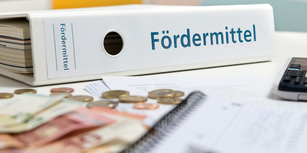 Auf einem Schreibtisch liegt ein Aktenordner mit der Aufschrift "Fördermittel", davor liegen Euro-Münzen und -Scheine.