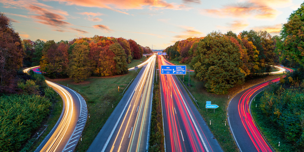 Eine deutsche Autobahn in der Abenddämmerung. Durch eine längere Belichtung bilden die Scheinwerfer und Rücklichter der Autos weiße und rote Linien auf der Fahrbahn.