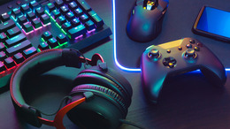 Das Bild zeigt ein Set aus Gaming-Zubehör auf einem Schreibtisch: eine beleuchtete mechanische Tastatur, eine Gaming-Maus auf einem leuchtenden Mauspad, ein schwarzer Gamecontroller, ein Smartphone und ein Paar Over-Ear-Kopfhörer. Alle Geräte sind in farbigem RGB-Licht inszeniert.