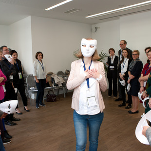 eine Person mit einer weißen Maske vor dem Gesicht, darum im Halbkreis weitere Personen
