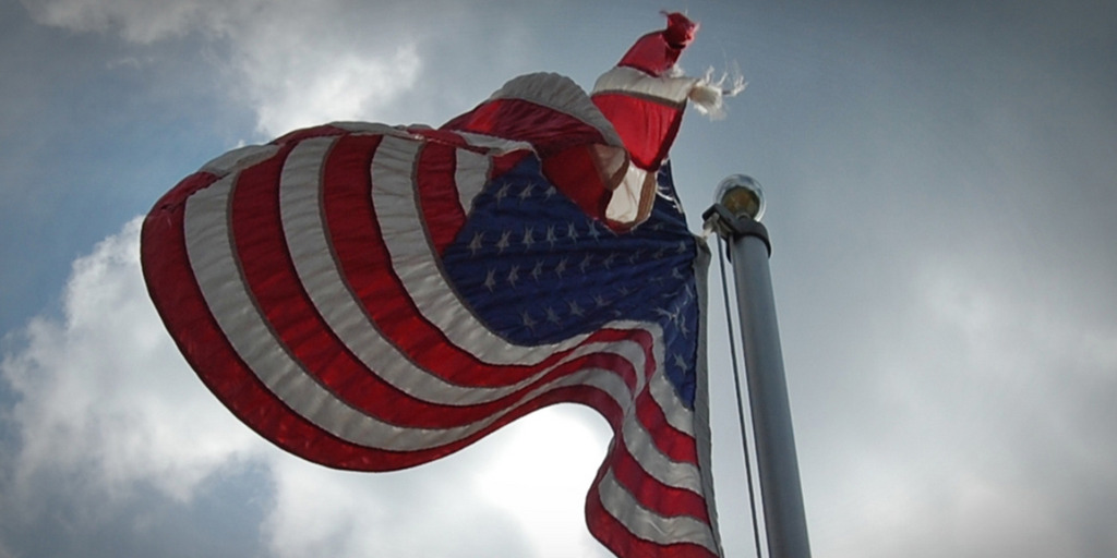 Vor einem bedrohlich dunkelgrau bewölkten Himmel weht eine leicht zerfledderte US-Flagge an einem Fahnenmast.