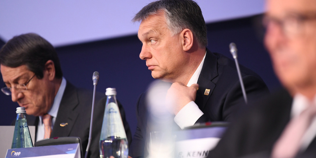 The Hungarian prime minister Viktor Orbán sits at a desk at a conference of the European People's Party. He is looking into the audience and touching his collar.