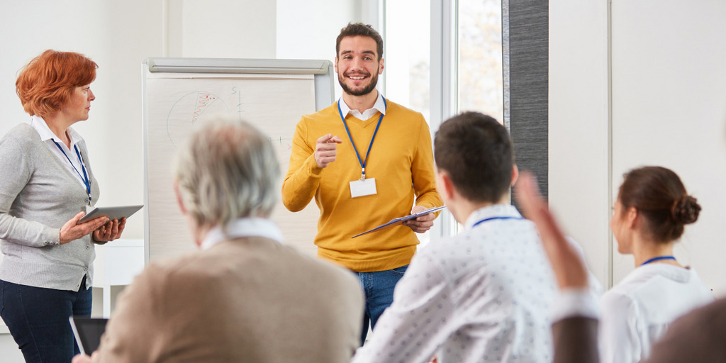 Gruppe Erwachsener unterschiedlichen Alters befindet sich in einer Schulung, einer junger Trainer steht vor einem Flipchart