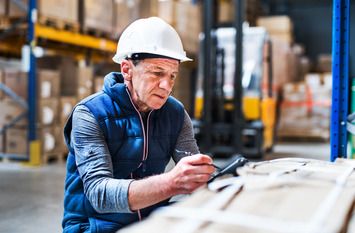 Ein älterer Herr mit Schutzhelm zeichnet an einem Werktisch in einer Lager- oder Fabrikhalle.