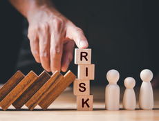 Eine menschliche Hand hält mit dem Zeigefiger vier Bauklötze fest, auf denen "RISK" geschrieben steht.