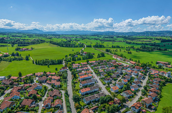 Das Bild zeigt eine ländliche Siedlung in Deutschland, eingebettet in eine weite, grüne Kulturlandschaft mit Wiesen, Feldern und vereinzelten Höfen. Im Hintergrund sind sanfte Hügel und Berge zu sehen.