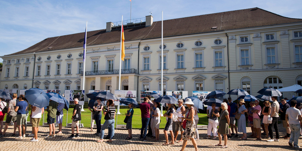 Vor dem Schloss Bellevue in Berlin stehen zahlreiche Menschen in einer Schlange. Viele von ihnen halten dunkelblaue Sonnenschirme in der Hand, um sich vor der Sonne zu schützen. Im Hintergrund ist die Fassade des Schlosses mit wehender Deutschlandfahne zu sehen.