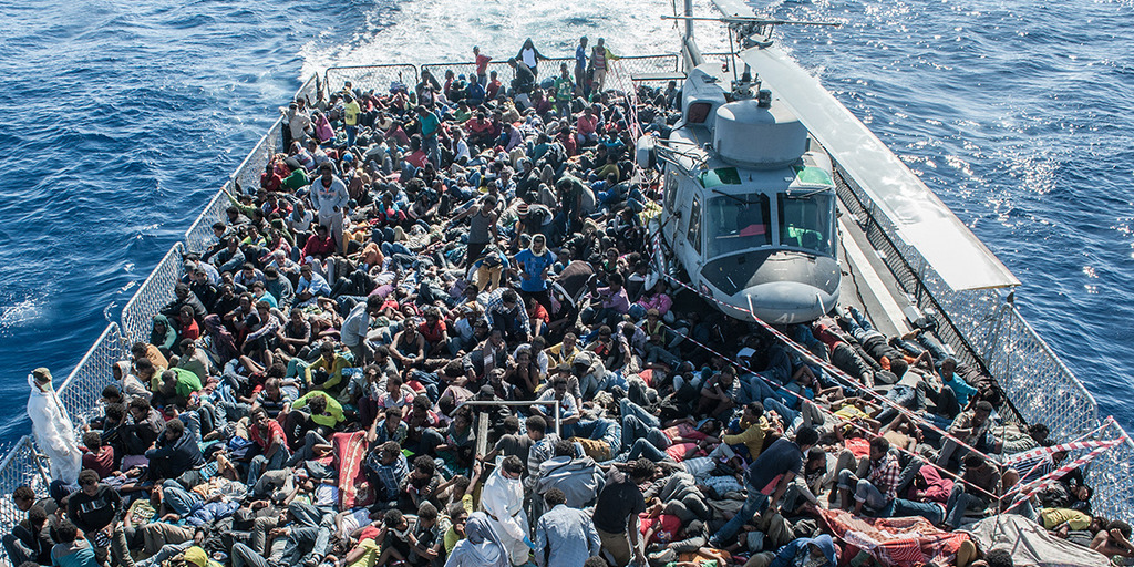 Vogelperspektive auf ein Rettungsboot voll mit Flüchtlingen und einem Hubschrauber im Mittelmeer.