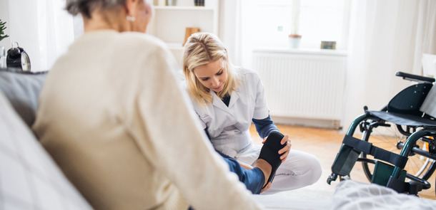 Eine junge Pflegekarft zieht einer älteren Frau zu Hause Pantoffeln an.