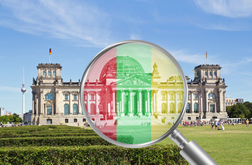 Das Bild zeigt das Berliner Reichstagsgebäude mit einer Lupe im Vordergrund, durch die das Gebäude in den Farben der Ampelkoalition (Rot, Gelb, Grün) hervorgehoben wird.