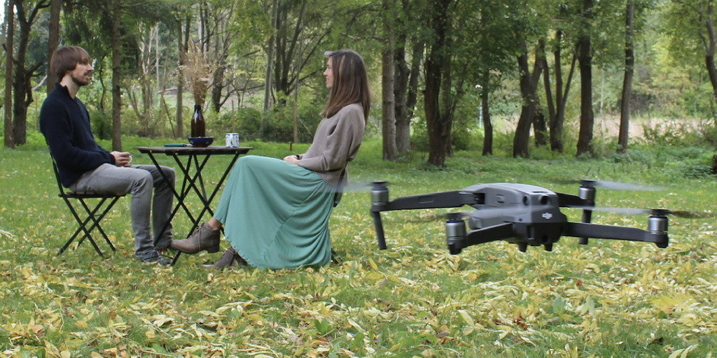 Mann und Frau sitzen im Wald gemeinsam an einem Tisch. Vor ihnen fliegt eine Drohne.