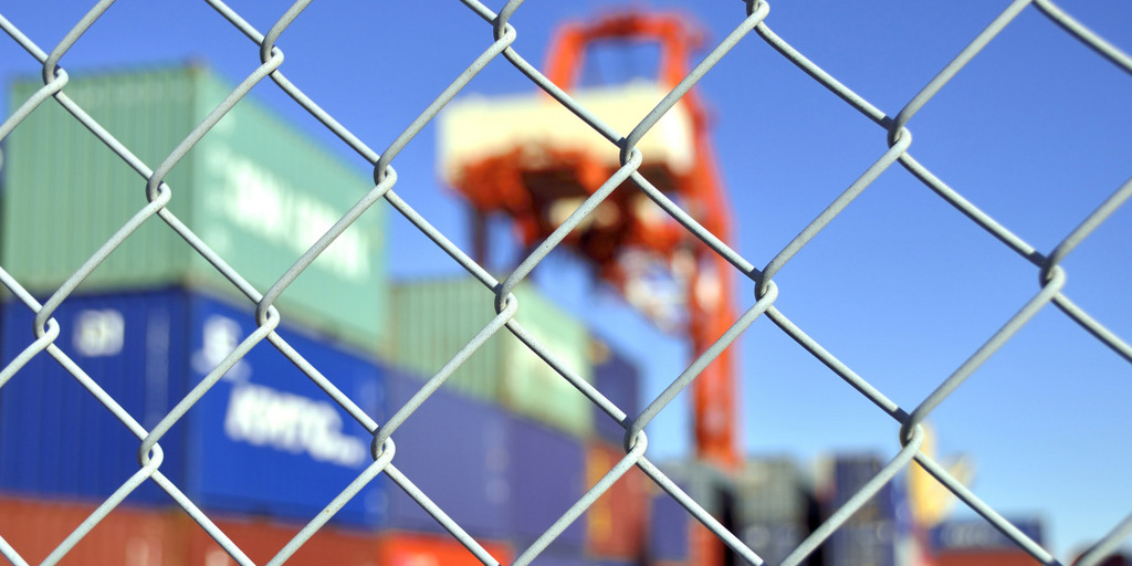A chain-link fence blocks the entrance to a harbor. A container crane and a few containers are visible in the background.