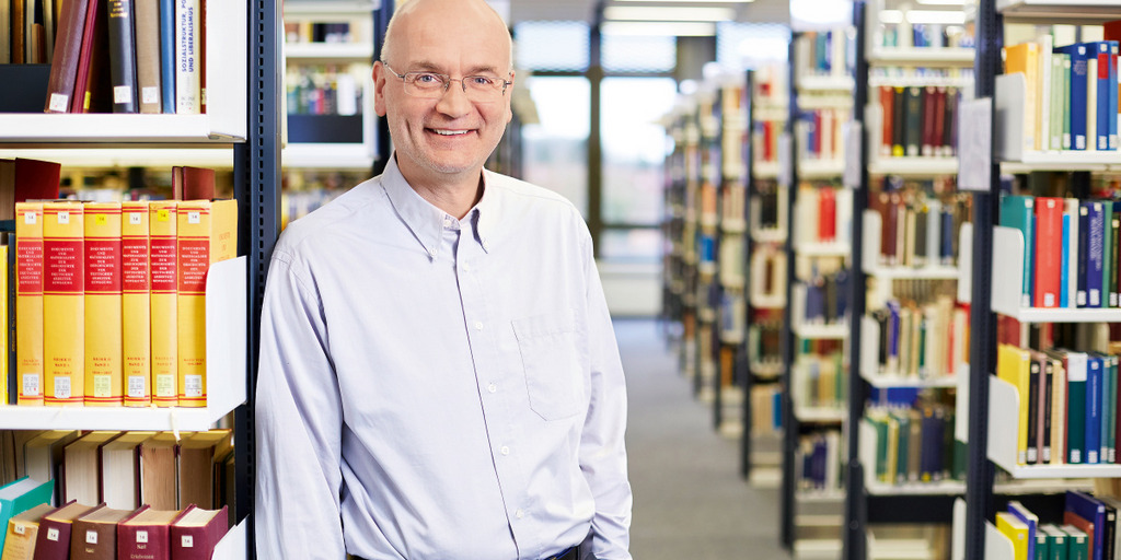 Prof. Udo Ohm steht vor einem Bücherregal in einem Raum der Universität Bielefeld.