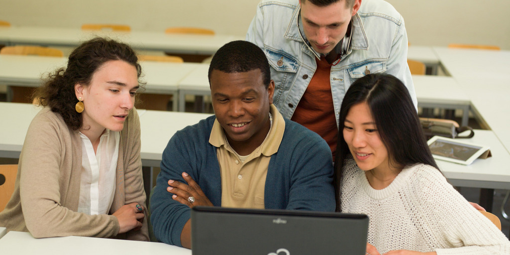 Zwei Studentinnen und zwei Studenten sitzen und stehen vor einem Laptop und besprechen etwas.