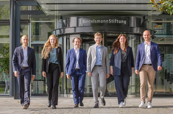 Die sechs Junior Professionals der Bertelsmann Stiftung des Jahrgangs 2023 - Jakob Weber, Anne Meisiek, Etienne Höra, Leander Berner, Lena Budach und Otto Meyer zu Schwabedissen - gehen gemeinsam den Weg vor dem Eingang zur Bertelsmann Stiftung hinunter und blicken in die Kamera.