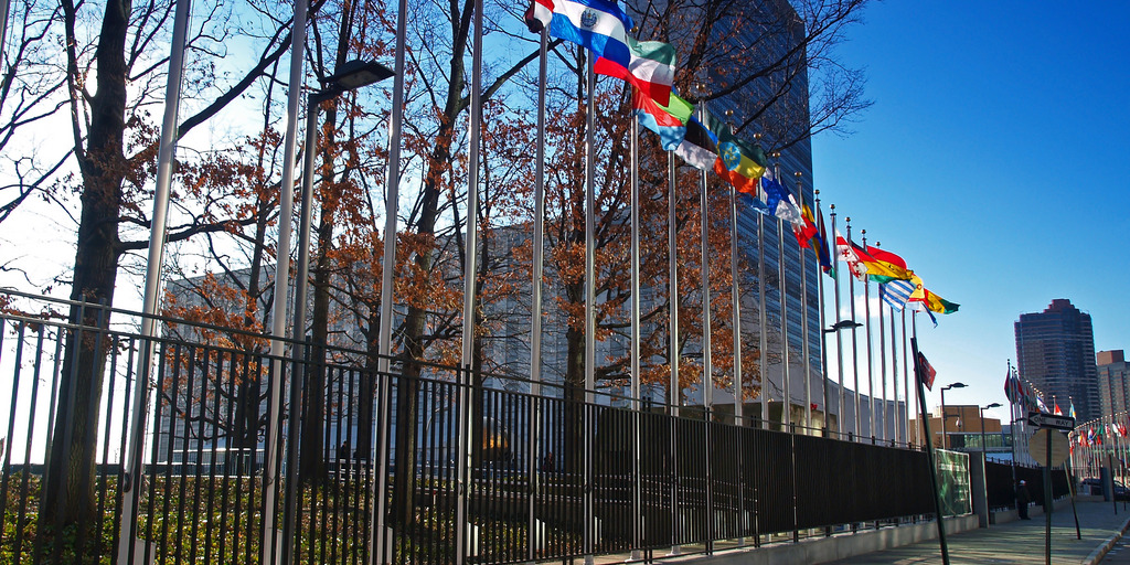 Zu sehen ist die Straße vor dem UN Gebäude. An der Straße stehen Fahnenstangen mit Fahnen der UN Staaten und ein paar Bäume