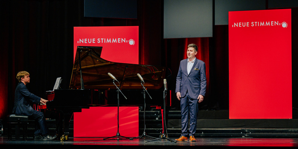Kieran Carrel, Teilnehmer des NEUE-STIMMEN-Wettbewerbs 2022, steht beim Vorsingen in der Endrunde in Gütersloh auf der Bühne und singt, neben ihm ist ein Pianist am Flügel zu sehen.