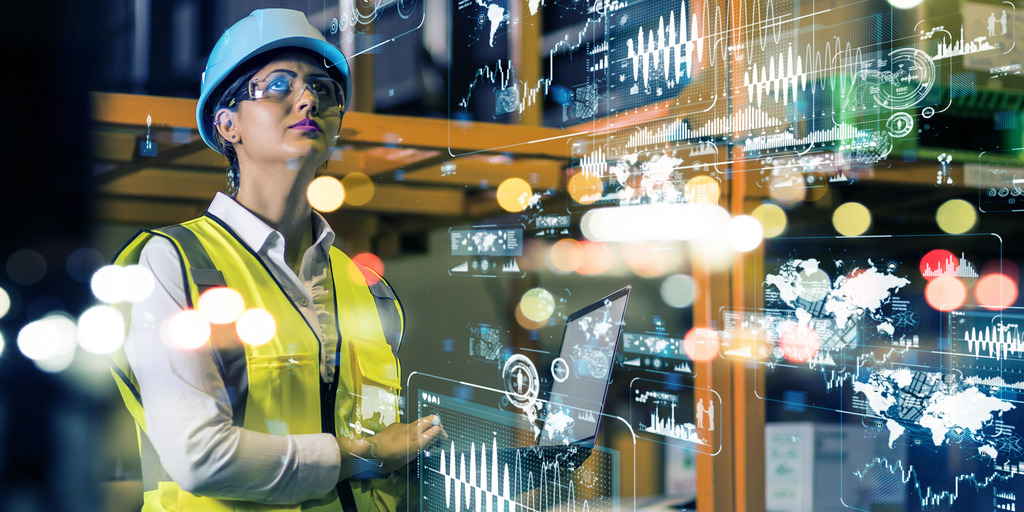 Eine junge Frau steht in einer Fabrikhalle, trägt Helm und gelbe Warnweste. Sie steht in einem Hologramm aus Lichtreflexen und Daten.