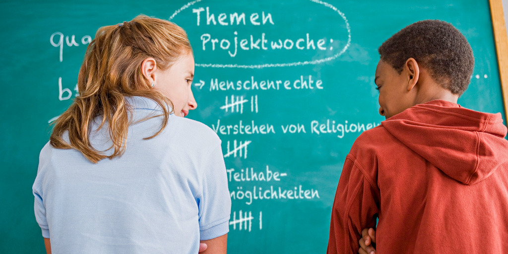 Ein Junge und ein Mädchen stehen mit dem Rücken zur Kamera vor einer Schultafel. An der Tafel stehen Begriffe wie "Menschenrechte" und "Teilhabemöglichkeiten"