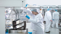 In einer Fabrikhalle arbeitet eine Frau mit Kittel, Haube und Handschuhen bekleidet an einer großen metallfarbenen Maschine zur Lebensmittelproduktion