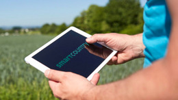 Person steht im Feld mit einem iPad. Der Oberkörper ist nur leicht zu sehen. Der Fokus liegt auf dem iPad