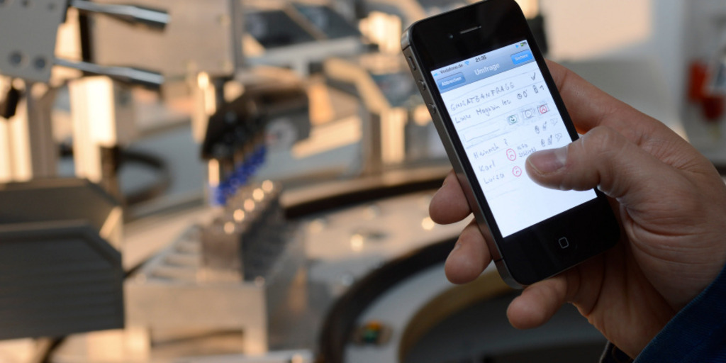 Ein Mann hält in einem produzierenden Unternehmen ein Smartphone in der Hand und bedient es. Im Hintergrund sind verschwommen technische Geräte.