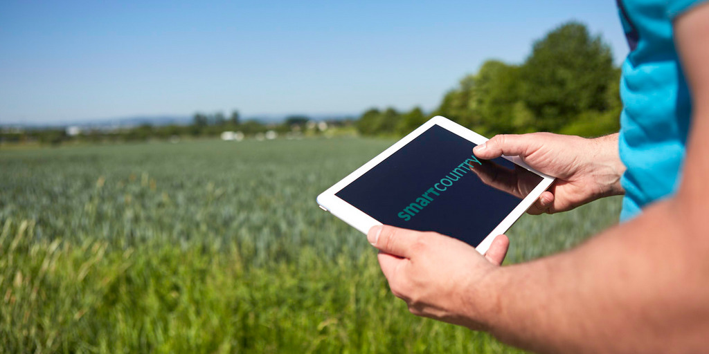 Eine Männerhand hält ein Tablet. Darauf steht der Schriftzug Smart Country. Im Hintergrund ist ein Maisfeld.