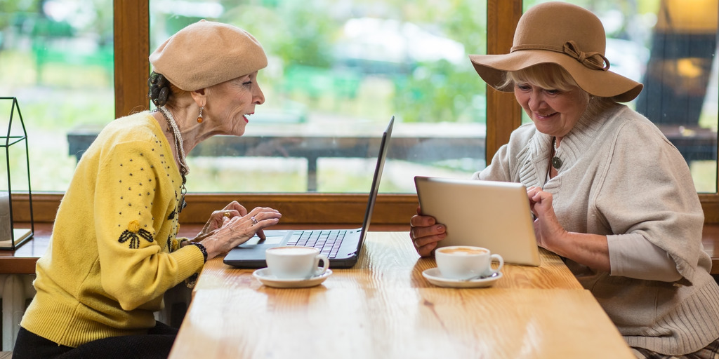 Zwei ältere Frauen sitzen am Tisch. Die eine Frau sitzt vor einem Laptop und schaut gespannt auf den Bildschirm. Die zweite Frau hat ein Tablet in der Hand und schaut auf dieses. Beide Frauen sehen glücklich aus uns schmunzeln.