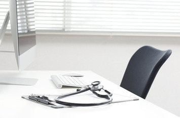 Helles, minimalistisches Arztzimmer: Auf einem weißen Schreibtisch liegen Stethoskop und Klemmbrett, daneben Tastatur, Maus und Monitor; im Hintergrund Jalousien und ein schwarzer Bürostuhl.