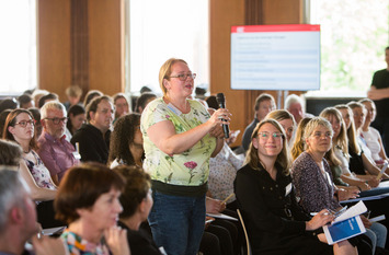 Eine Teilnehmerin steht bei einem Treffen des Bürgerrates für das Forum gegen Fakes im Saal und spricht in ein Mikrofon, während ihr andere Teilnehmer:innen zuhören.