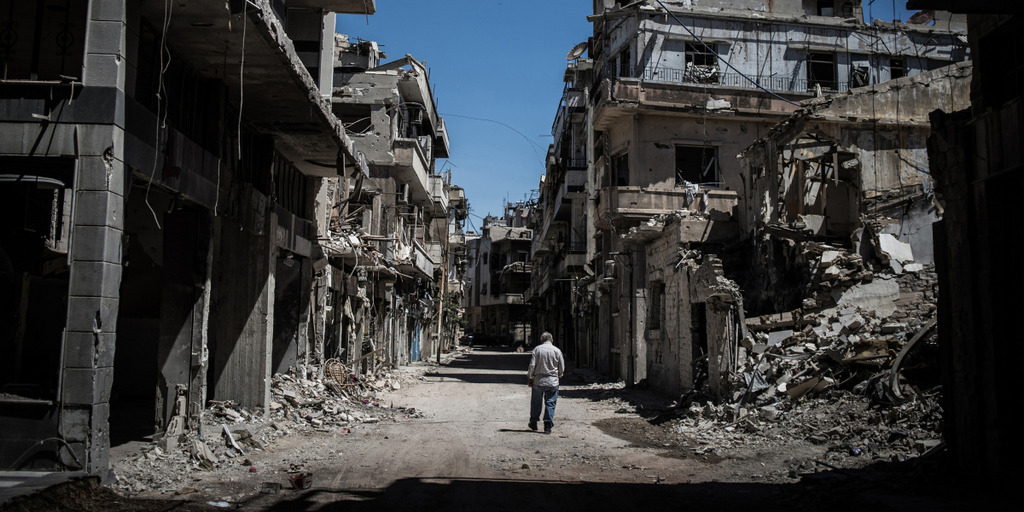 Ein Mann läuft eine Straße im syrischen Homs entlang. Links und rechts stehen die Ruinen von fast vollständig zerstörten Häusern.