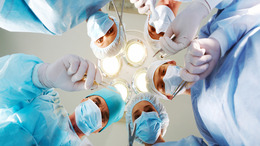 Aus der Perspektive eines im Operationssaal liegenden Patienten sind fünf Ärzte in ihren Operationskitteln und mit Mundschutz zu sehen, die sich über den Patienten beugen und Scheren und Skalpelle in der Hand haben. Sie blicken dem Patienten in die Augen.