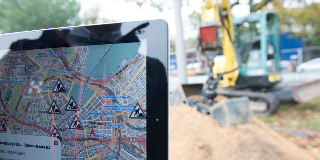 Eine nicht sichtbare Person hält ein Tablet in die Kamera, auf der ein Stadtplan von Hamburg mit eingezeichneten Baustellen-Standorten zu sehen ist. Im Hintergrund ist verschwommen eine Baustelle mit einem Bagger auf einem Sandhaufen zu erkennen.