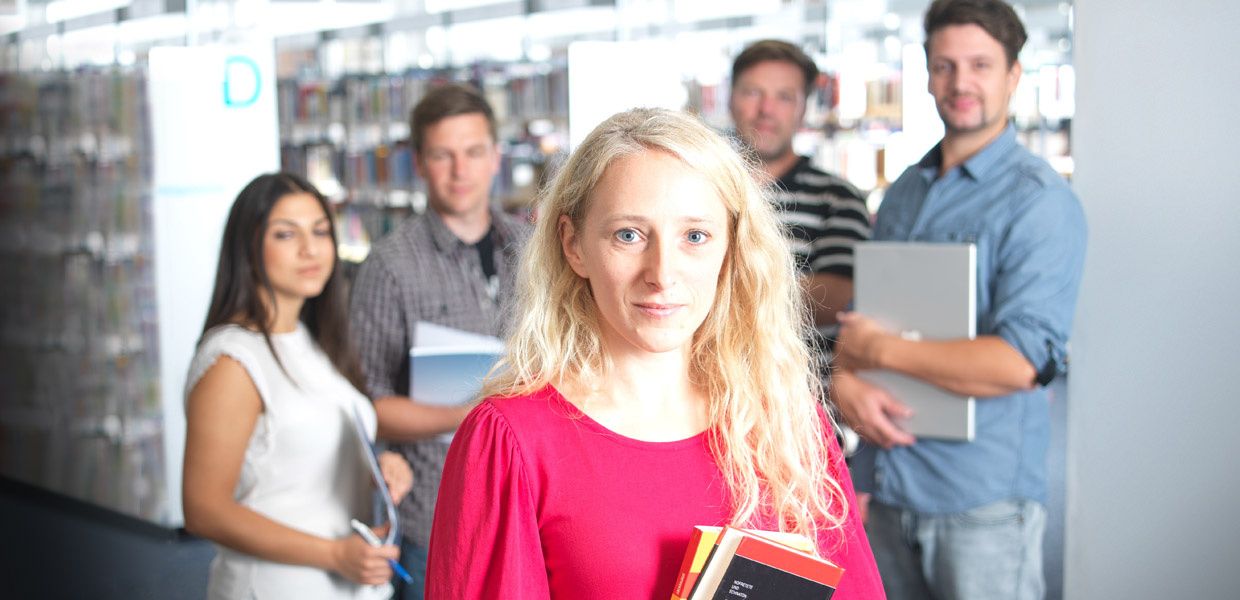 (© Arne weychardt / Bertelsmann-Sti) Eine Frau mit Broschpren in der Hand steht im Vordergrund. Im Hintergrund sind im Halbkreis 4 weitere Personen und eine Bibliothek von innen schemenhaft zuerkennen.