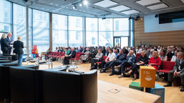 Der Blick geht vom Podium aus in den Konferenzraum. Der Raum ist hell mit Fensterfront vom Boden bis zur Decke. Auf den Stühlen sitzen in Reihen die Teilnehmer.