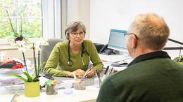 Das Bild zeigt eine freundliche Szene in einer Arztpraxis: Eine Ärztin im Gespräch mit einem älteren Patienten. Beide sitzen sich an einem Schreibtisch gegenüber. Die Ärztin trägt ein Stethoskop um den Hals und hört aufmerksam zu. Im Hintergrund ist ein Computerbildschirm zu sehen, auf dem medizinische Daten angezeigt werden.
